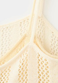 Gros plan d'un tissu tricoté couleur crème avec des motifs en maille ouverte et de fines bretelles, montrant la texture et les détails des points.