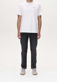 Hombre vestido con una camiseta de manga corta blanca de GABBA, jeans negros y zapatillas gris claro, de pie contra un fondo blanco liso.