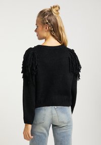 Pulover negru din tricot, cu accente texturate de franjuri pe umeri, guler rotund și lungime scurtă. Purtați cu blugi albaștri deschis.