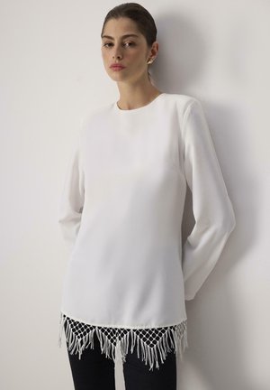 White long-sleeve top with a round neckline, featuring a fringe hem with netted detailing. Smooth fabric, tailored fit, and straight silhouette.