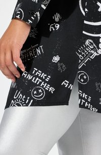 Top de tela negra con texto gráfico blanco y símbolos. Cuenta con un puño abotonado y un dobladillo en ángulo. Combinado con leggings plateados brillantes.