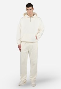 Cremefarvet hættetrøje med halvlukning, stor lomme og ribbede manchetter, parret med matchende joggers. Glat, blød tekstur hele vejen igennem.
