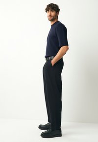 Tumši zils īsroku džemperveida krekls kopā ar melnām biksēm ar krokām un melniem apaviem. Modelis pozē ar rokām kabatās uz vienkārša fona.