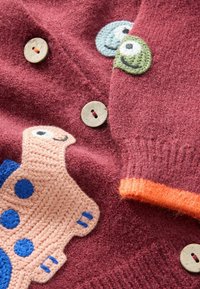 Primer plano de tela de punto burdeos con botones de madera y parches tejidos de colores con forma de animales con caras sonrientes.