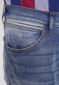 Área del bolsillo de unos pantalones vaqueros de mezclilla azul con costuras naranjas y un ligero desgaste, usados sobre una camisa a rayas roja, blanca y azul.
