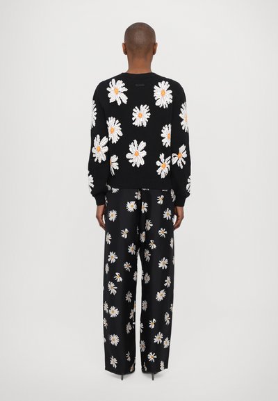 Schwarzer Pullover mit einem weißen und orangefarbenen Gänseblümchenmuster; kombiniert mit schwarzen weit geschnittenen Hosen, die dasselbe florale Design aufweisen. Gerippte Bündchen und Saum.