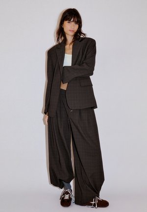Kvinde poserer i oversize mørkt ternet jakkesæt med brede bukser, hvid cropped top, grå sokker og brune sneakers mod en ensfarvet baggrund.