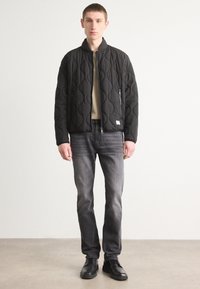 Black quilted bomber jacket with a stand collar, front zipper, and two side pockets, paired with gray jeans and black shoes on a light background.