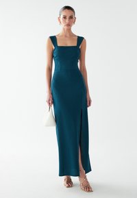 Blågrön ribbad maxiklänning med fyrkantig urringning, breda band och sidslits. Matchas med guldfärgade remsandaler och en vit handväska.