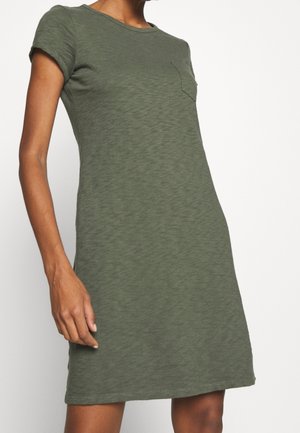 Mujer que lleva un vestido casual de manga corta, verde oliva, hasta la rodilla, con un pequeño bolsillo en el pecho a la izquierda.