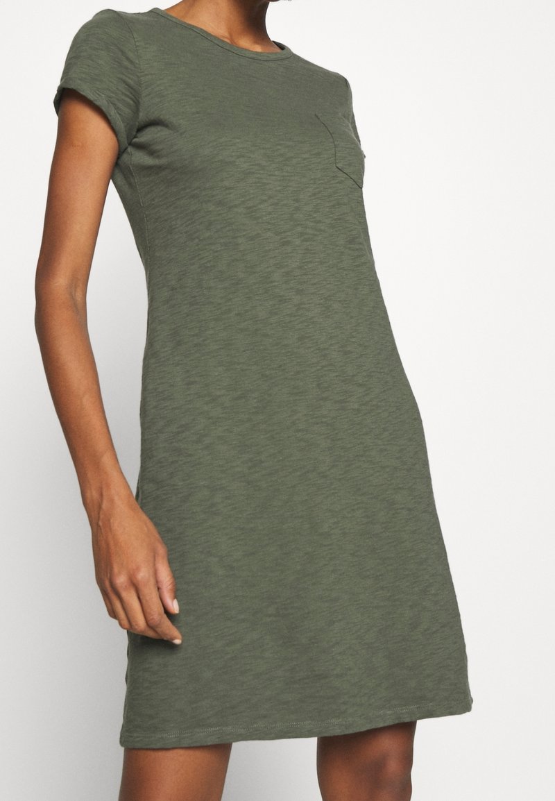 Femme portant une robe décontractée courte manches, vert olive, longueur genou, avec une petite poche poitrine sur le côté gauche.