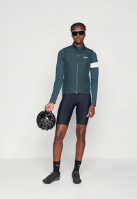 Herenfietsvest in donker teal met witte accenten, gemaakt van lichtgewicht materiaal. Model draagt zwarte shorts en houdt een zwarte helm vast.