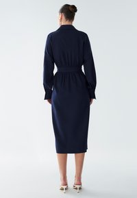 Abito midi blu navy con maniche lunghe a sbuffo, vita stretta e silhouette dritta, realizzato in tessuto liscio. Indossato con tacchi bianchi.