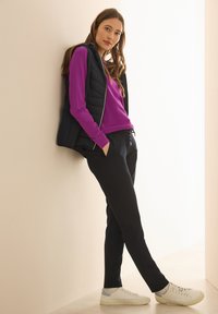 Gilet matelassé navy sur un pull violet, associé à un pantalon noir et des baskets blanches. La tenue présente une texture lisse et un design décontracté.