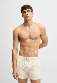 Men's swim shorts in light beige fabric with a textured pattern, elastic waistband, and drawstring for adjustability.