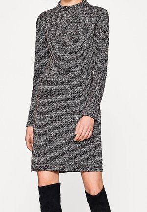 Kobieta w teksturowanej, dzianinowej sukience z długim rękawem do kolan, w kolorze szaro-czarnym, zestawiona z czarnymi, sięgającymi do kolan wysokimi butami.