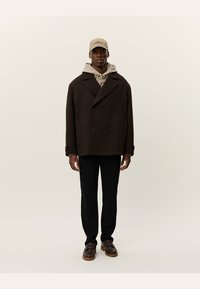 Abrigo marrón de doble botonadura con grandes botones, llevado sobre una sudadera beige con capucha. Combinado con pantalones negros y zapatos marrones. Gorra en color beige claro.