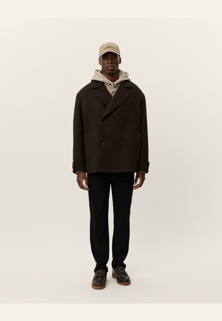 Abrigo marrón de doble botonadura con grandes botones, llevado sobre una sudadera beige con capucha. Combinado con pantalones negros y zapatos marrones. Gorra en color beige claro.