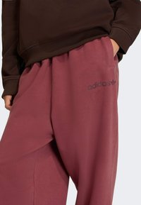 Pantalones de chándal color marrón de adidas con una cinturilla elástica, que cuentan con bolsillos laterales y un pequeño logo negro en el muslo izquierdo. Textura suave.