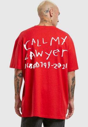 Rød bomulds t-shirt med oversized pasform, der har håndskrevne hvide bogstaver: "KALD MIN ADVOKAT" og numre på ryggen. Korte ærmer, rund halsudskæring.