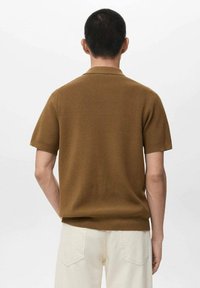 Polo marrone a maniche corte realizzata in tessuto lavorato a maglia, con colletto classico e vestibilità morbida, indossata con pantaloni di colore chiaro.