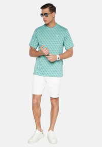 T-shirt verde menta con un motivo ripetuto, abbinato a pantaloni corti bianchi e sneaker bianchi. Accessoriato con occhiali da sole grigi e un orologio bianco.