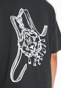 Sort t-shirt med et stort hvidt grafisk motiv af et dekorativt emblem med indviklet linjearbejde og et stiliseret bogstav "M."