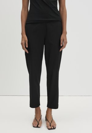 Donna con pantaloni neri aderenti alla caviglia e sandali neri con tacco e cinturini, in piedi davanti a uno sfondo chiaro e uniforme.