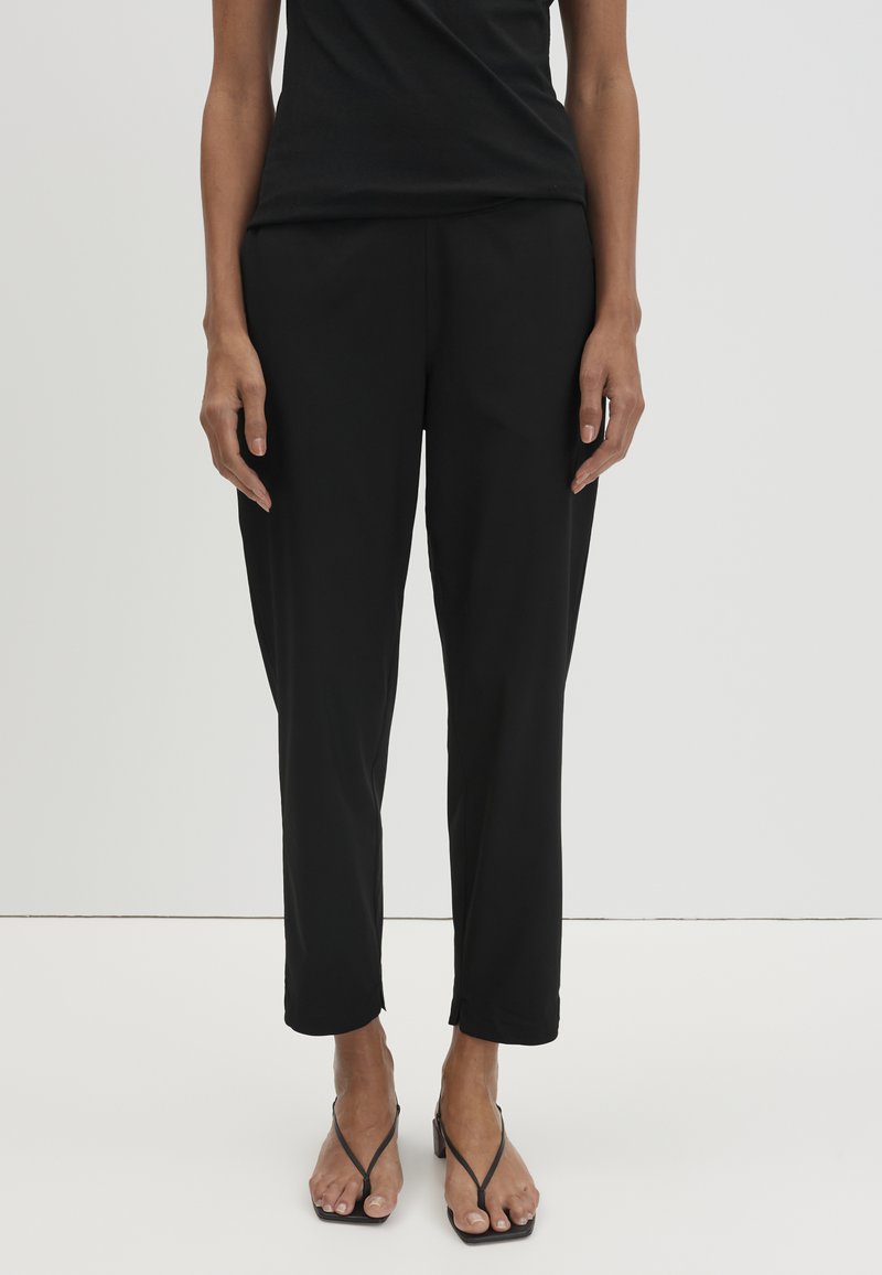 Vrouw met zwarte taps toelopende enkellange broek en zwarte hakken met bandjes, staand tegen een effen lichte achtergrond.