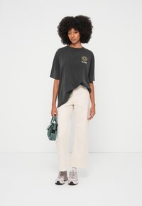 Szary, oversize'owy t-shirt z krótkimi rękawami, z grafiką słońca. W połączeniu z kremowymi spodnicami o szerokich nogawkach i jasnoszarymi sneakersami, trzymający turkusową torbę.