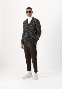 Grå cardigan med diamantmønster, mørkebrun cordbukser og hvite sneakers. Modell som har på seg solbriller og en hvit høyhalset genser.