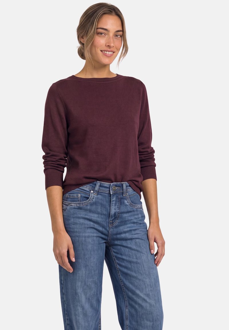 Jeune femme aux cheveux châtain clair en chignon, portant un pull bordeaux à manches longues et un jean bleu, souriante et debout devant un fond uni.