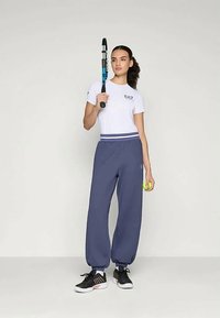 Λευκό t-shirt με λογότυπο EA7, ναυτικές μπλε φόρμες, μαύρα αθλητικά παπούτσια, κρατώντας μια ρακέτα και μια μπάλα τένις, σε ουδέτερο φόντο.