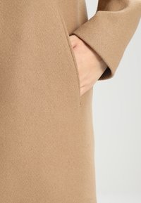 Cappotto beige con tasca e design elegante; il tessuto ha una texture liscia con una finitura leggermente strutturata e bordi puliti.