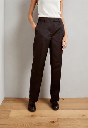 Pantalon noir sur mesure avec design à jambe droite, texture lisse et plis marqués, associé à des chaussures noires sur un sol en bois.