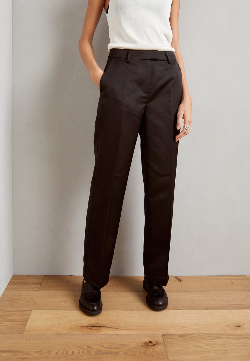 Black tailored trousers with a straight-leg design, smooth texture, and pressed creases, paired with black shoes on wooden floor.