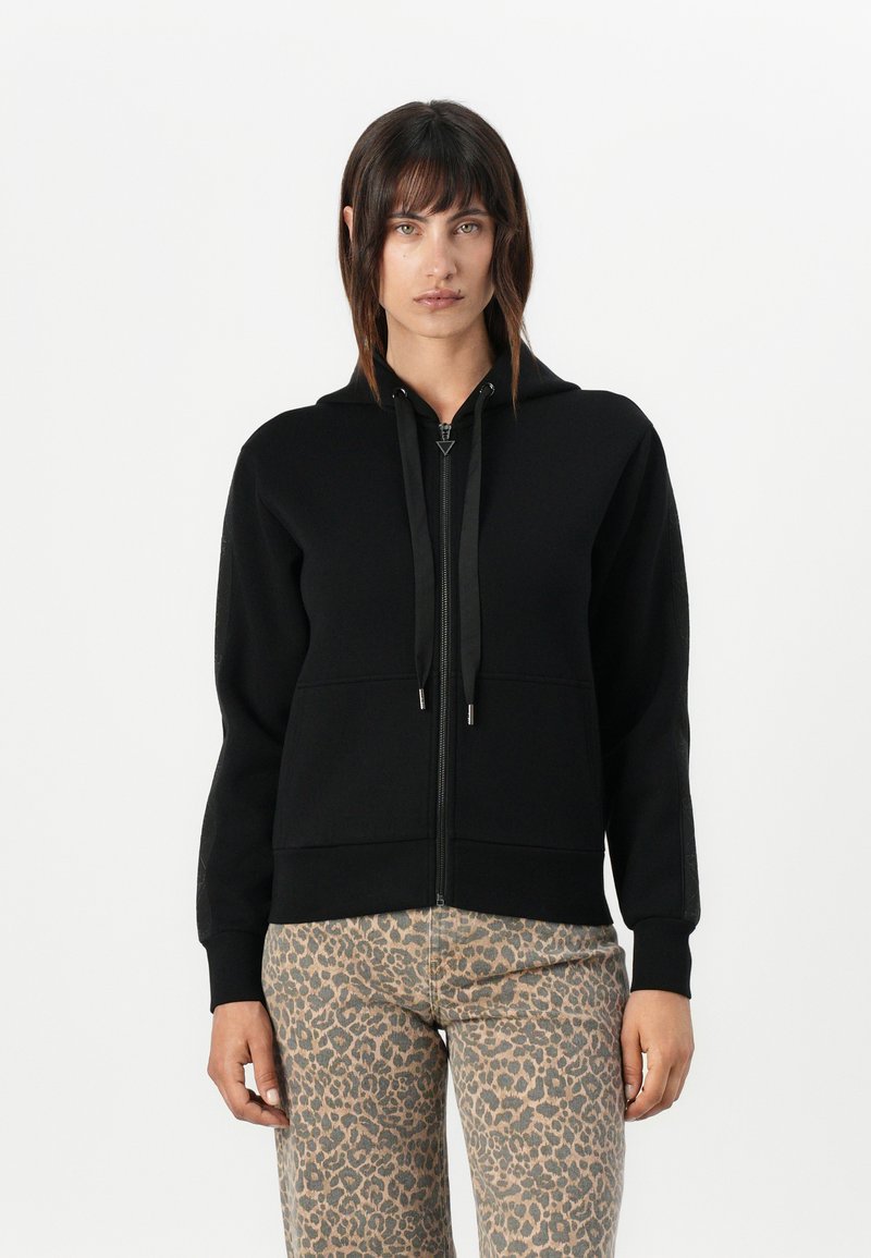 Zwarte zip-up hoodie met een capuchon met trekkoord, voorzien van een gladde textuur en voorzakken, gecombineerd met beige broek met luipaardprint.