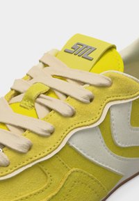 Baskets jaunes et blanches avec lacets beige, textures en suède et cuir, et un logo gris sur la languette.