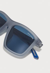 Ochelari de soare cu un cadru din plastic gri mat, lentile de culoare albastru închis și un accent albastru pe brăcuș. Prezintă o formă pătrată și branding minimal.