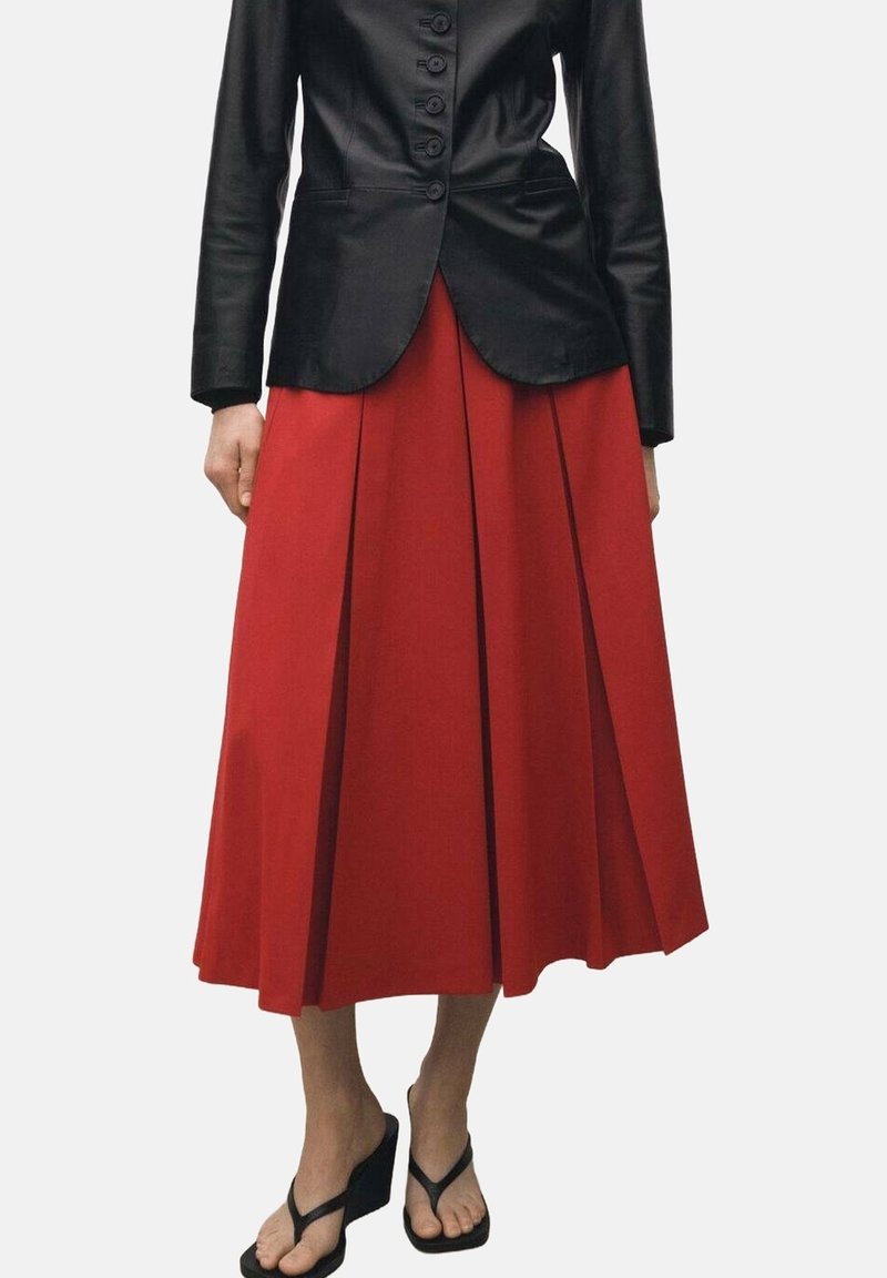 Femme portant une veste noire cintrée boutonnée, une jupe midi plissée rouge et des sandales noires compensées à brides, debout devant un fond uni.