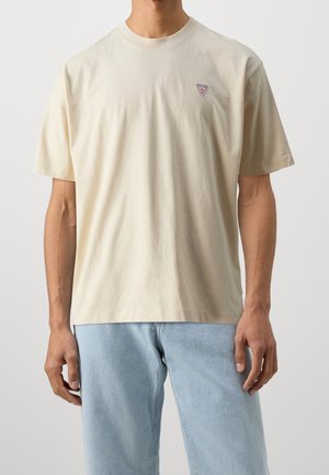 Hombre vistiendo una camiseta beige lisa con un pequeño logo triangular de Guess en el pecho y jeans azul claro.