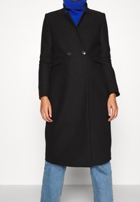 Abrigo negro a medida con botones de doble pecho, mangas largas y bolsillos laterales. Lleva puesto un jersey de cuello alto azul y lo combina con jeans claros.
