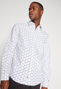 Camicetta bianca con pattern di testo nero che legge "TOMMY JEANS." Presenta un colletto puntato e maniche lunghe. Tessuto di cotone liscio.