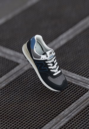 Zwarte en grijze sneaker met witte veters en accenten. Blauwe hiel, gestructureerd materiaal en een duidelijke "N"-logo aan de zijkant. Rubberen zool.