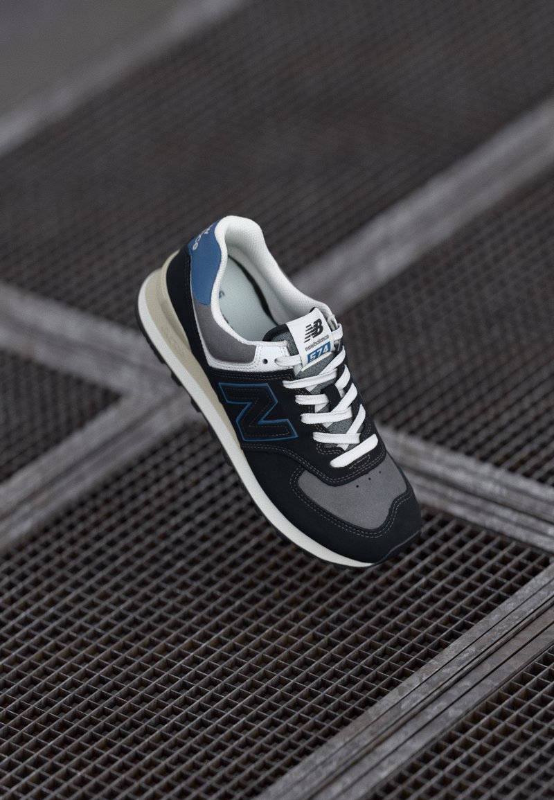 Black and gray sneaker with white laces and accents. Blue heel, textured material, and distinct "N" logo on the side. Rubber outsole.