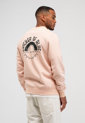 Man draagt lichtroze sweatshirt met zwart "Butcher of Blue" logo en tekst op de rug, crèmekleurige broek, staat in profiel tegen effen achtergrond.