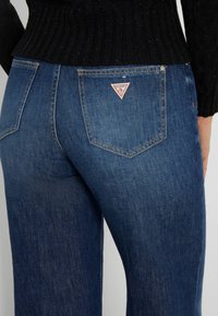Jeans de mezclilla con un lavado azul oscuro, que presentan un parche de logo triangular en el bolsillo trasero, y un diseño de pierna recta con un desvanecimiento mínimo.