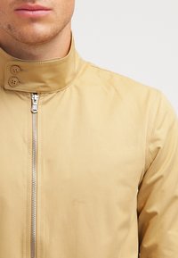 Beige letvægtsjakke med høj krave, der har en frontlynlås og to knaplukninger på kraven. Glat, mat tekstur.