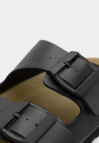 Schwarze Ledersandale mit zwei verstellbaren Riemen, ausgestattet mit mattschwarzen Schnallen und einer strukturierten Innensohle auf einem hellbraunen Fußbett.