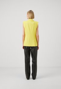 Femeie cu păr scurt blond, purtând un blazer galben fără mâneci și pantaloni din piele negri, stând cu spatele împotriva unui fundal simplu.